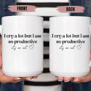 May include: Two white ceramic mugs with black text that reads "I cry a lot but I am so productive it's an art" and a small heart symbol. The mugs are held by hands.