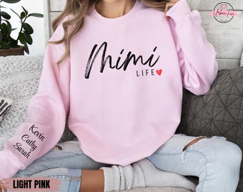 May include: Pale pink sweatshirt featuring the text "Mimi LIFE" in black script with a red heart. The left sleeve displays the names "Kevin, Cathy, Sarah" in black. A comfortable, casual style.