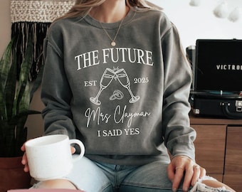 May include: A gray sweatshirt with the text "The Future Est 2025" and "Mrs. Clayman I Said Yes" in white lettering. The sweatshirt features a graphic of two champagne glasses clinking with a ring in between.
