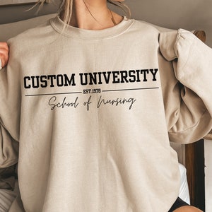 Könnte beinhalten: Ein beiger Pullover mit dem Aufdruck "CUSTOM UNIVERSITY EST. 1978 School of Nursing" auf der Vorderseite.