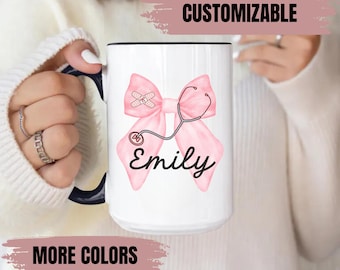 May include: White ceramic mug with a black handle and rim. The mug features a pink bow design with a stethoscope and a bandage, along with the name "Emily" in a script font. The words "CUSTOMIZABLE" and "MORE COLORS" are also visible.