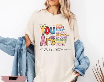 May include: Ivory t-shirt with a colorful graphic that says "You Are" with a list of positive words like "Kind, Brave, Smart, Helpful, Grateful, Enough, Strong, Unique, Loved". The text "Mrs. Emma" is below the graphic.