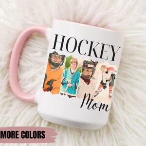 Puede incluir: Taza de cer&aacute;mica blanca con asa rosa. La taza presenta las palabras "HOCKEY Mom" en texto negro, junto con cuatro im&aacute;genes de jugadores de hockey. Las palabras "MORE COLORS" est&aacute;n en la parte inferior de la taza.