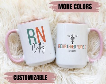 May include: Two white ceramic mugs with pink handles. One mug has the letters RN in colorful block letters with the name Cathy written below. The other mug has a black and white caduceus symbol with the text "Registered Nurse Est. 2025".