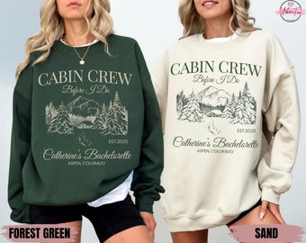 May include: Forest green and sand-coloured sweatshirts with the text "CABIN CREW Before I Do" and "Catherine's Bachelorette Aspen, Colorado". The design includes a mountain scene with trees and a cabin. The sweatshirts are long-sleeved.