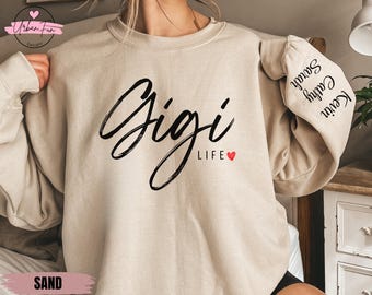 May include: A sand-coloured sweatshirt featuring the words "Gigi LIFE" in black script, a small red heart, and names on the sleeve. The sweatshirt has a crew neck and long sleeves. The Urban Fun Designs logo is in the upper left corner.