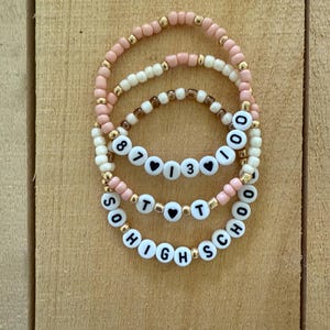 May include: Three beaded bracelets with white letter beads spelling out "8713", "TOT", and "SOHIGHSCHOOL". The bracelets feature pink, cream, and brown beads with gold accents. The bracelets are displayed on a wooden surface.