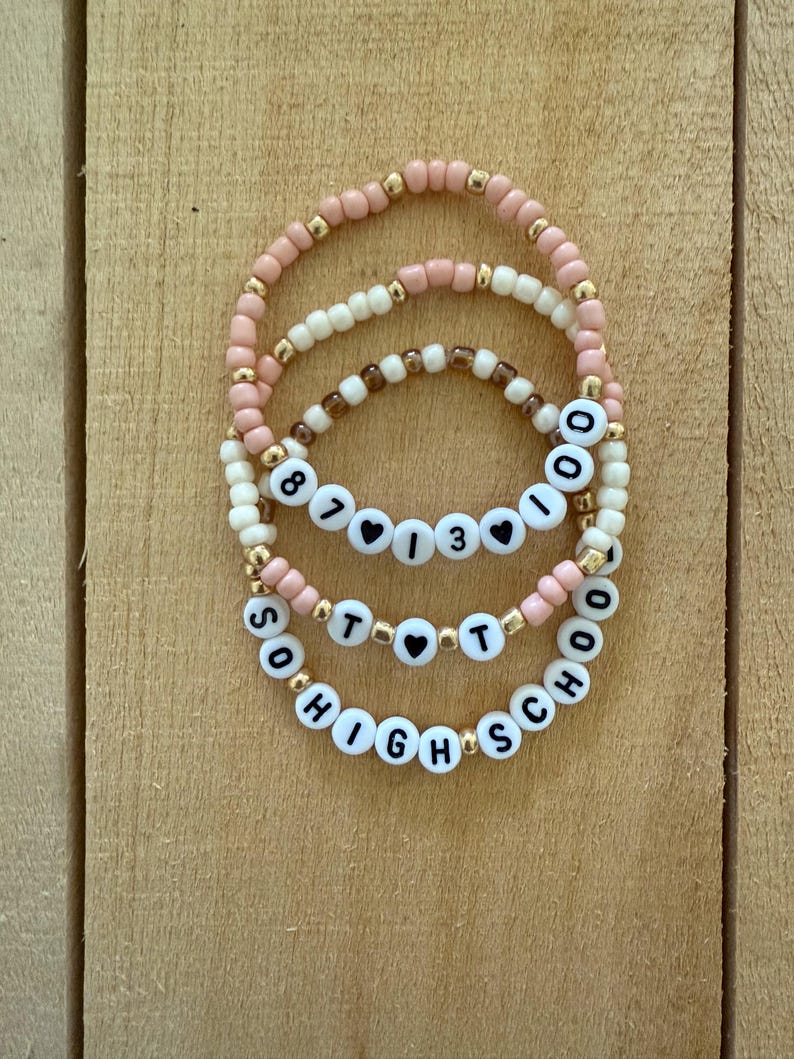 May include: Three beaded bracelets with white letter beads spelling out "8713", "TOT", and "SOHIGHSCHOOL". The bracelets feature pink, cream, and brown beads with gold accents. The bracelets are displayed on a wooden surface.