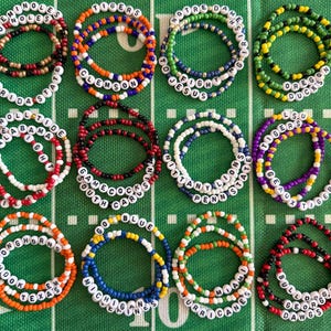 May include: A collection of beaded bracelets with team names, including "Seminoles," "Tigers," and "Longhorns." The bracelets feature various colors and white letter beads, arranged on a green football field-themed background.