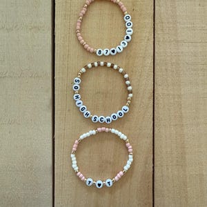 May include: Three beaded bracelets with white letter beads spelling out words and numbers. The bracelets are in shades of pink, white, and gold. The bracelets are on a light brown wooden surface.