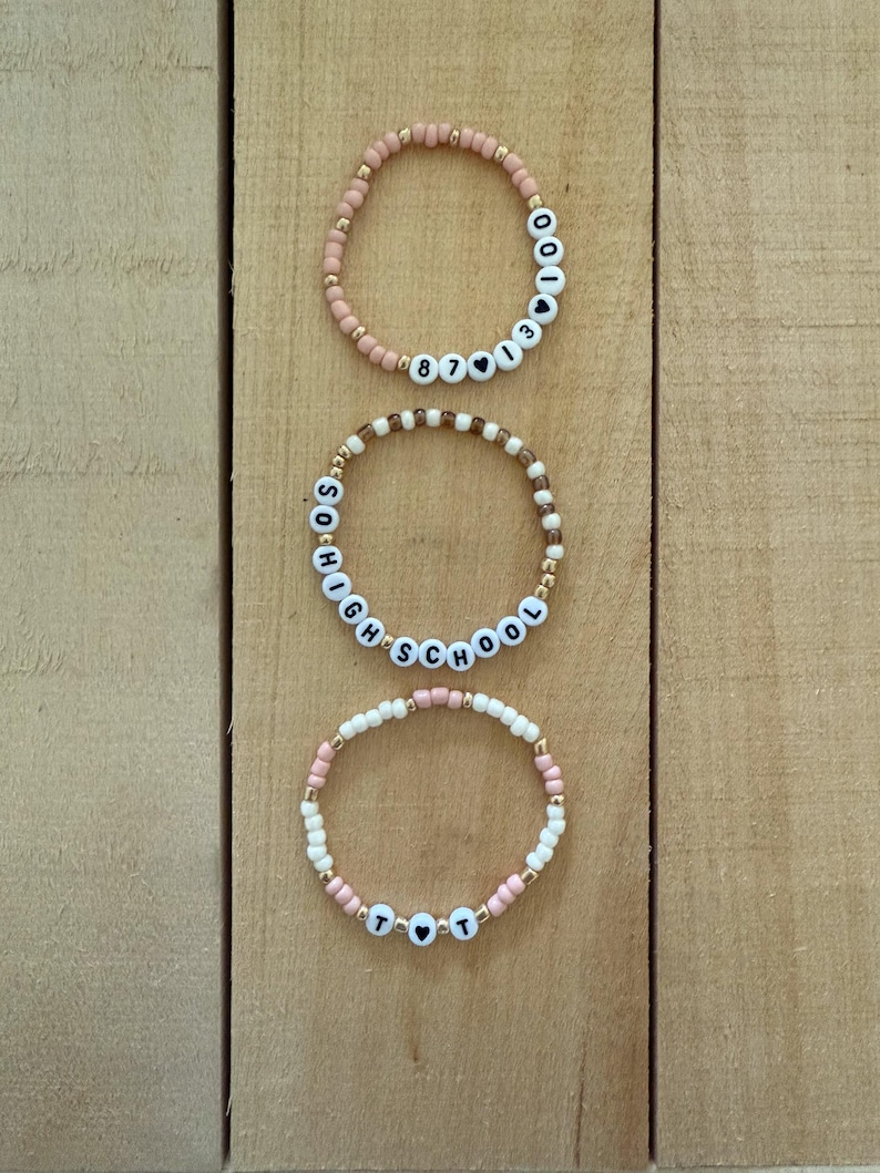 May include: Three beaded bracelets with white letter beads spelling out words and numbers. The bracelets are in shades of pink, white, and gold. The bracelets are on a light brown wooden surface.