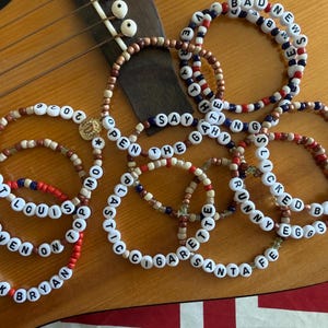 May include: A collection of beaded bracelets with white letter beads spelling out phrases like "Zack Bryan," "Heaven on Top," and "Bad News." The bracelets feature red, white, and blue beads, with a gold charm.