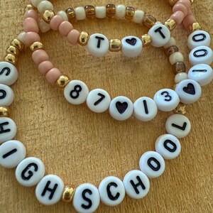 May include: Three beaded bracelets with white letter beads spelling out "HIGH SCHOOL" and "87 I 13". The bracelets feature pink, cream, and gold beads. The bracelets are on a wooden surface.