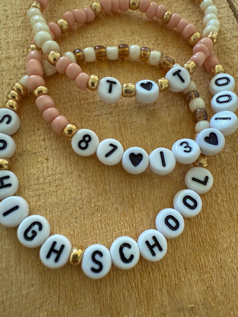 May include: Three beaded bracelets with white letter beads spelling out "HIGH SCHOOL" and "87 I 13". The bracelets feature pink, cream, and gold beads. The bracelets are on a wooden surface.