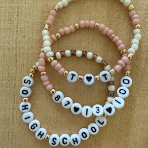 Könnte beinhalten: Drei Armbänder aus Perlen in Rosa, Creme und Gold. Die Armbänder haben weiße Buchstabenperlen mit der Aufschrift "HIGH SCHOOL", "87", "13" und "TOO". Herzförmige Perlen sind ebenfalls enthalten.