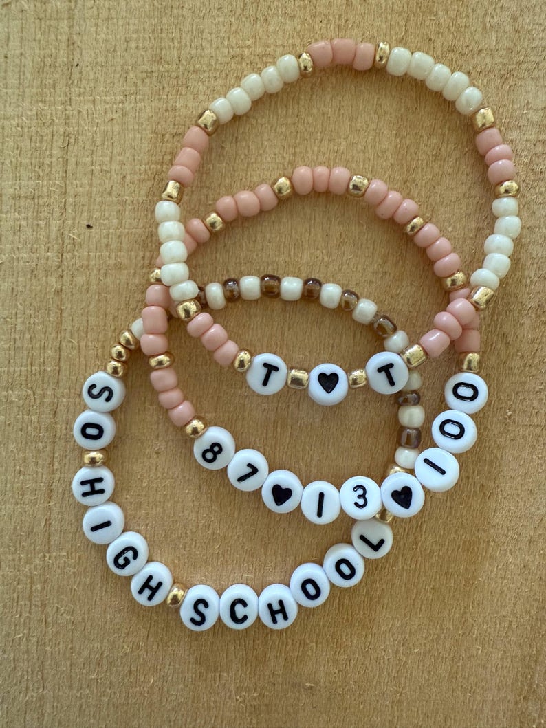 May include: Three beaded bracelets in shades of pink, cream, and gold. The bracelets feature white letter beads spelling out "HIGH SCHOOL", "87", "13", and "TOO". Heart-shaped beads are also included.