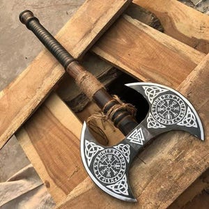 Op de afbeelding: Een dubbelzijdige strijdbijl met een donkerhouten steel omwikkeld met natuurlijk jutegaren. De metalen bijlkop is voorzien van gedetailleerde, wit gegraveerde Noorse symbolen, waaronder het Vegvisir-kompas en Valknut-knooppatronen.