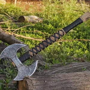 Op de afbeelding: Een dubbelbladige bijl met een zilverkleurige metalen kop met ingewikkelde gegraveerde patronen. De steel is gemaakt van hout en is omwikkeld met zwart leren koord in een kruispatroon, met extra zwarte leren omwikkeling bij de handgreep.