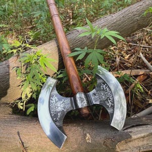 Könnte beinhalten: Eine Doppelaxt mit einem langen, glatten, braunen Holzstiel und einem breiten Kopf aus poliertem Stahl. Die Metallklinge ist auf beiden Seiten mit detaillierten Gravuren von Fabelwesen und verschlungenen Knotenmustern verziert.