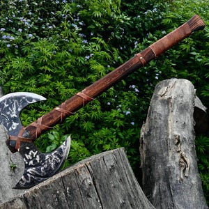May include: A double-bit axe featuring a dark, textured metal head with a weathered finish and a long, polished wooden handle. The handle is wrapped with thin, brown leather cord detailing that spirals along the shaft and secures the axe head.