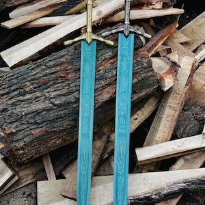 Op de afbeelding: Twee zwaarden met lichtblauwe bladen voorzien van een golvend patroon. Het linkerzwaard heeft een goudkleurig gevest en pareerstang, terwijl het rechterzwaard een zilverkleurig gevest en pareerstang heeft. Beide bladen zijn recht en puntig.