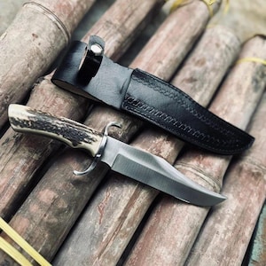 Peut inclure: Un couteau à lame fixe avec un manche en bois de cerf texturé, une garde en métal argenté et une lame en acier poli. Le couteau est accompagné d'un étui en cuir noir doté d'une fermeture à bouton-pression et de coutures décoratives.