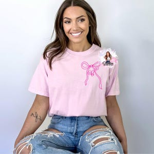 May include: A pink t-shirt with a pink bow graphic on the front. The shirt is being worn by a person with long brown hair.