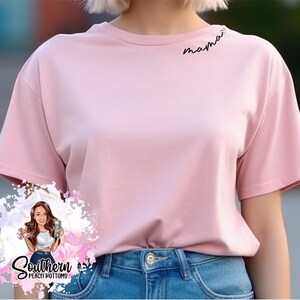 Op de afbeelding: Een roze t-shirt met korte mouwen met het woord "mama" geborduurd op de voorkant in zwarte cursieve letters.