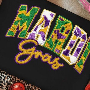 May include: Black fabric with the embroidered words "Mardi Gras" in yellow, green, purple, and white. The letters have a textured, patch-like appearance. The image is decorated with Christmas ornaments and a leopard print.