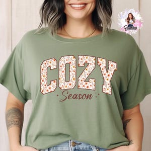 May include: Olive green t-shirt with the word "COZY" in a floral design, and the word "Season" below. The shirt has short sleeves and a relaxed fit, perfect for casual wear.