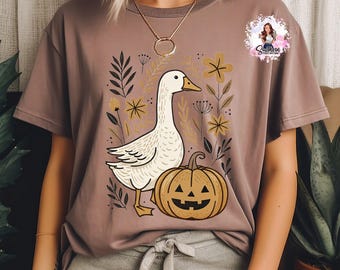 May include: A mauve-colored t-shirt featuring a whimsical illustration of a white goose, a jack-o'-lantern, and floral elements. The design has a fall or Halloween theme, with a cartoonish style. The shirt is a casual wear item.