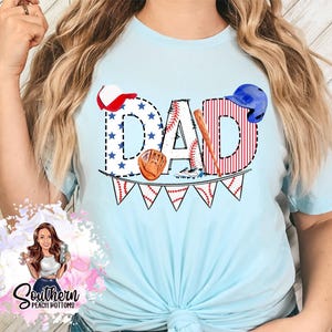 May include: A light blue t-shirt featuring a graphic design of the word "DAD" in red, white and blue with baseball-themed imagery, including a baseball bat, glove and baseballs.