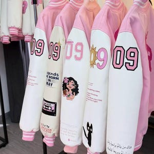 Peut inclure: Plusieurs vestes de style universitaire roses et blanches suspendues. Les vestes ont des manches et des cols roses, avec des corps blancs affichant le numéro "09" et divers graphiques et textes. Les poignets et les bas sont rayés rose et blanc.