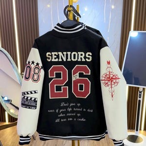 May include: Black varsity jacket with white sleeves. The back features "SENIORS 26" in red and white lettering. The sleeves have the number "08" and a compass rose design. The jacket has a striped collar and cuffs, with the text "Don't give up even if your life turned to dust when mixed up, I'll turn into a cookie."