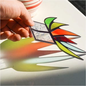 May include: A stained glass suncatcher shaped like a stylised window logo with a trailing flame design. The piece features clear textured glass panels and vibrant segments of green, yellow, orange, red, and light blue glass with black lead lines.