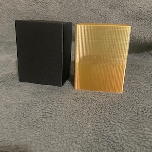 May include: Two rectangular boxes, one black and one gold, sit side-by-side. The boxes have a textured, ridged surface. The black box is matte, while the gold box has a metallic sheen. The boxes are on a gray, textured surface.