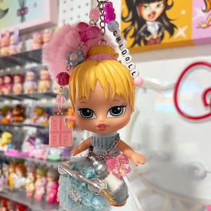 Puede incluir: Un llavero con una muñeca de pelo rubio y ojos azules grandes. La muñeca lleva un top gris y una falda azul con un lazo rosa. El llavero incluye un pompón rosa, cuentas y un dije que dice "CHIDOLL".