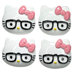 Può includere: Quattro decorazioni bianche a forma di viso di Hello Kitty. Ognuna ha occhiali con montatura nera, un fiocco rosa e un naso giallo. Le decorazioni sono disposte a quadrato su sfondo bianco.