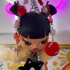 Puede incluir: Un llavero de muñeca Bratz con cabello negro, labios rojos y un cuello de piel sintética amarilla. El llavero presenta un cierre plateado, cuentas y el nombre "ASHLEY". También se adjuntan un dije de gato negro y un colgante de cruz.