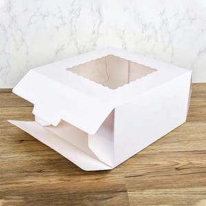 Può includere: Scatola per torte in cartone bianco con una finestra trasparente sulla parte superiore. La scatola è aperta e il coperchio è ripiegato all'indietro.