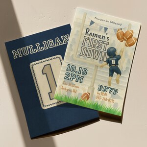 Può includere: Due inviti. Uno è blu navy con "MULLIGAN" e il numero 1. L'altro è un invito a tema football con il testo "Roman's FIRST DOWN" e un'illustrazione di un giocatore di football. Include data, ora e informazioni RSVP.