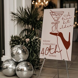 Puede incluir: Un cartel blanco con una ilustración roja de una copa de martini y un tacón alto. El cartel dice "A tiny bit older" y "Welcome to Samantha's 40th Birthday".