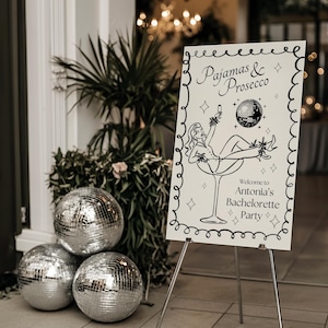 May include: A black and white sign with the text "Pajamas & Prosecco" and a graphic of a woman sitting in a martini glass. The sign is on a silver easel and there are three disco balls in the foreground.