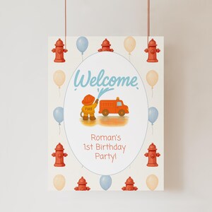 May include: Welcome to Roman's 1st Birthday Party! A welcome sign featuring a cartoon firefighter spraying water from a hose onto a red fire engine. The sign is decorated with red fire hydrants and blue and white balloons.