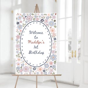 May include: A white welcome sign with a floral border and a white oval in the center. The oval has the text "Welcome to Madelyn's 1st Birthday" in black and brown lettering.