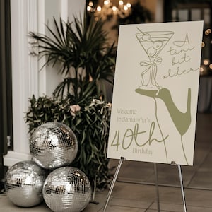 May include: A beige sign with a green illustration of a high heel holding a martini glass with olives. The sign reads "A tint bit older" and "Welcome to Samantha's 40th Birthday".