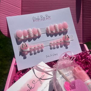 May include: A pink box contains press-on nails, nail glue, and an alcohol pad. The nails are pink and white with a French tip design. The card reads "Nails By Bri Press Ons" and "Thank you for supporting my small business."