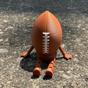 May include: A brown football-shaped figurine with white stitching detail. The figurine has articulated legs and arms, giving it a seated pose. The object is made of a 3D-printed material.