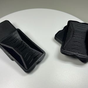 May include: Two black, 3D-printed phone cases. One case is open, revealing the interior design, while the other is closed. The cases have a textured surface and a unique shape, likely designed for a specific phone model.