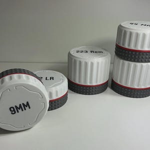 May include: A collection of four cylindrical white containers with ribbed sides and a textured dark gray base. Each lid features black printed text including 9MM, 22 LR, 223 Rem, and 44 MAG. A thin red ring separates the white top from the gray base.